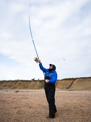 Barnaby in blue jacket and black pants casting a PENN Tidal Elite fishing rod on a beach.