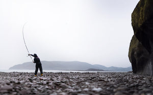 Person casting a fishing lure from a rocky shoreline with mountains in the background