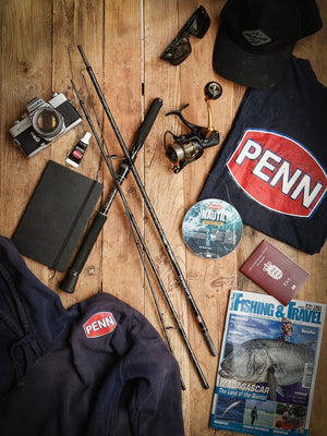 A selection of fishing gear and accessories as if just unpacked from a holiday bag  including a PENN travel rod, spinning reel, a camera, notebook, and a magazine on a wooden surface with a Penn branded hoodie and shirt.