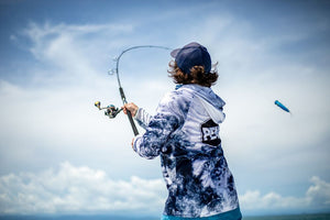 A person from behind wearing a blue and white tie-dye hoodie with a part visible PENN logo on the back, casting a fishing rod under high flex, set against a blue sky with clouds.