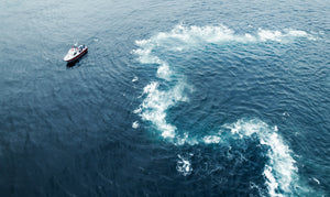 A high drone shot of a small white boat in the near still ocean. The blue ocean is only broken by a small white surf from an underwater crop. 