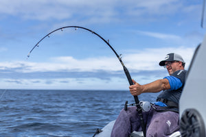 Man fishing on a boat with a fishing rod curved under the high tension of a catch against a blue sky and ocean.