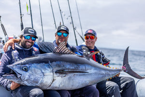 Three anglers in PENN caps and sunglasses resting a large tuna across their legs. With multiple rods and the ocean in the background